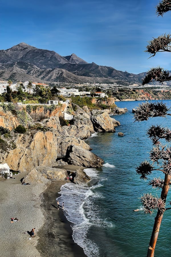 A gray sandy beach with deep blue water and rocky cliffs, and mountains in the distance.