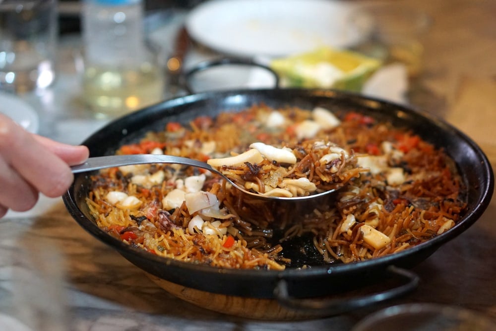 Close-up of fideuá, a noodle dish served in a large pan, with someone scooping a spoonful.