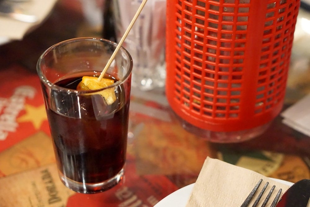 A glass of dark brown vermouth with ice and a slice of orange on a wooden skewer.