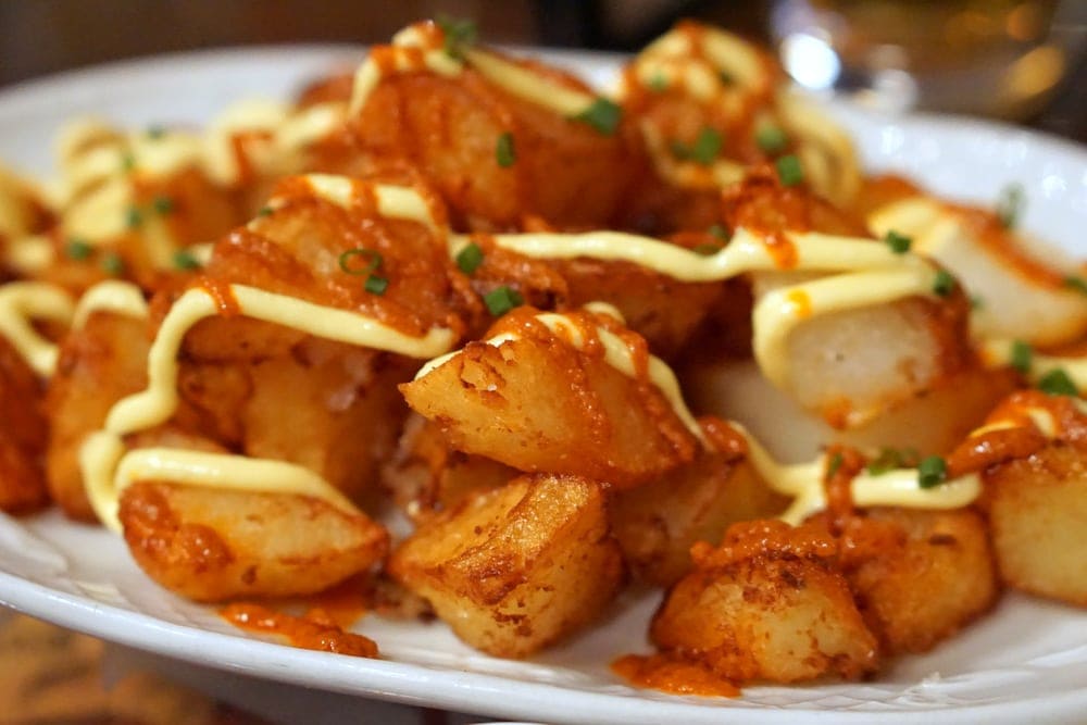 Close-up of a plate of fried potato chunks with orange bravas sauce, alioli, and chives.