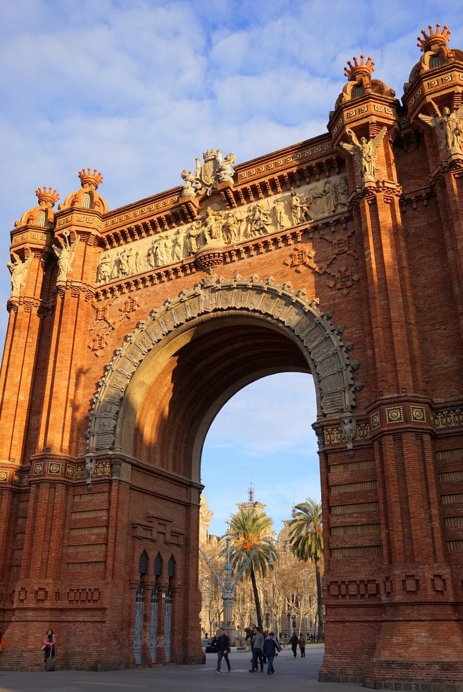 A large red brick arch covered in statues and mosaics, with people walking beneath it.