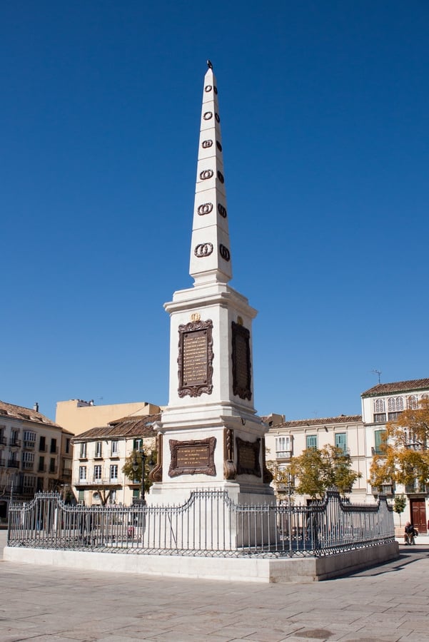 Tall obelisk made of white stone with dark brown plaques, in the center of a square.