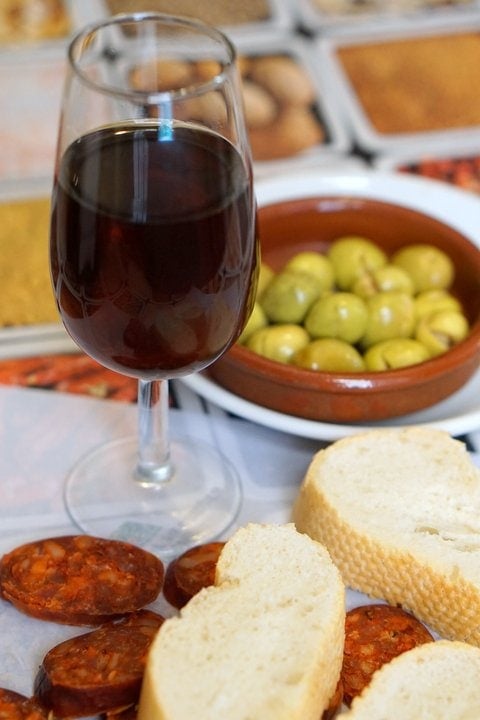 A table with a glass of sherry wine, bread, chorizo, and olives.