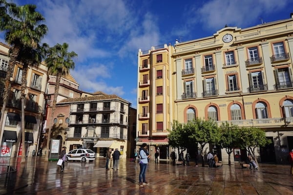 A historic square with a large yellow building on one side and tall skinny palm trees.