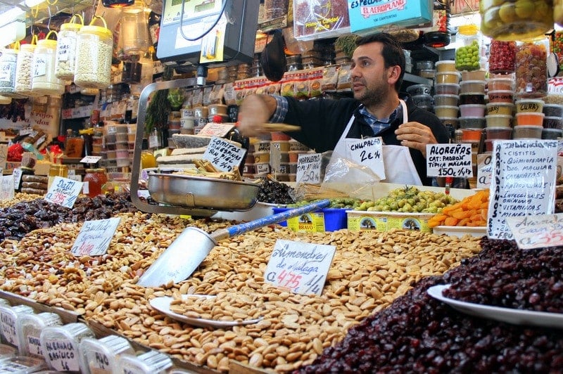 A market stall with bulk nuts, dried fruits, and spices, with the vendor standing behind it.