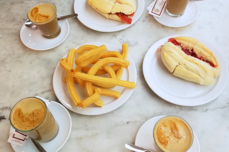 A table with four coffees, two sandwiches, and a plate of churros, seen from above.