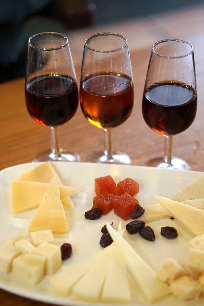 White plate with five different types of cheese, raisins, quince jelly, and three glasses of sherry.