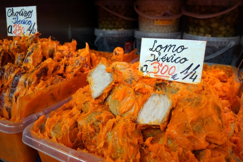 Large container of pork pieces coated in bright orange lard, with a sign reading "lomo ibérico."