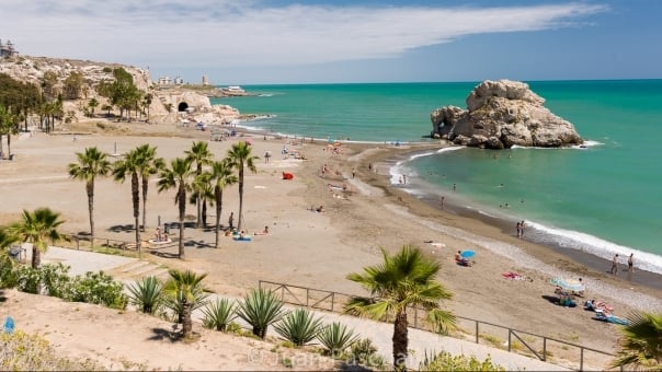 Wide view of a sandy beach with palm trees, turquoise water, and a large rock offshore.