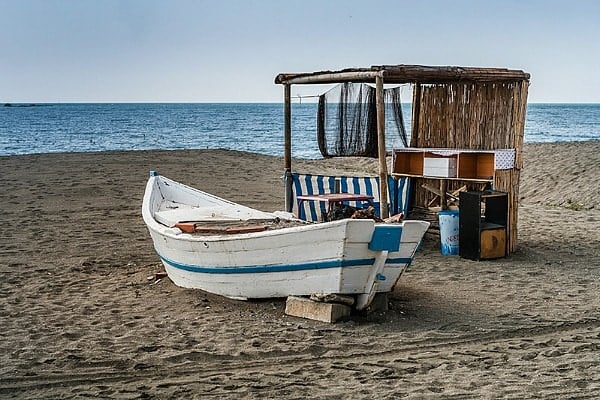 A sandy beach with a small wooden shelter and old white rowboat with a blue stripe.