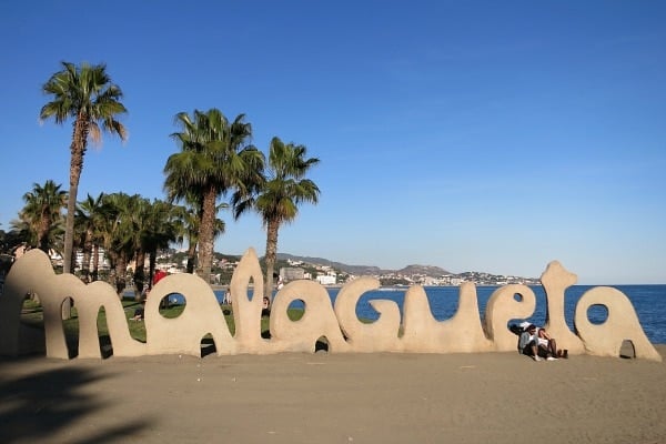 Large beige letters spelling out "Malagueta" with the ocean and palm trees behind them.