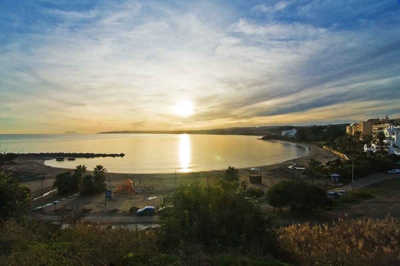 Wide view of a circular bay at sunset, with a sandy beach and some buildings nearby.