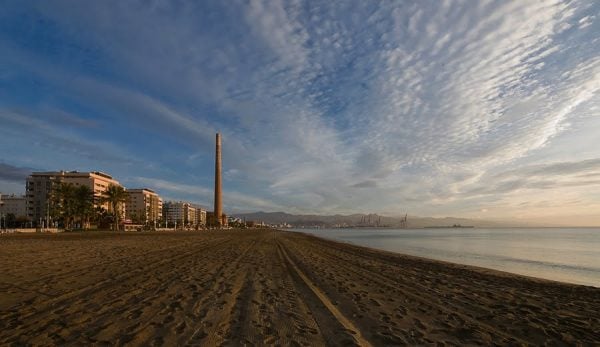 A sandy beach at sunset, with a tall skinny chimney rising up in the distance.