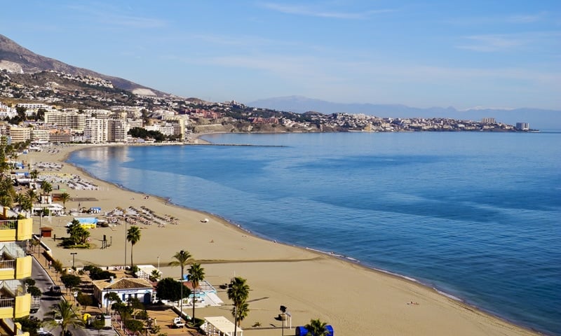 Wide view of a sandy beach with calm blue water and a city right beside it.