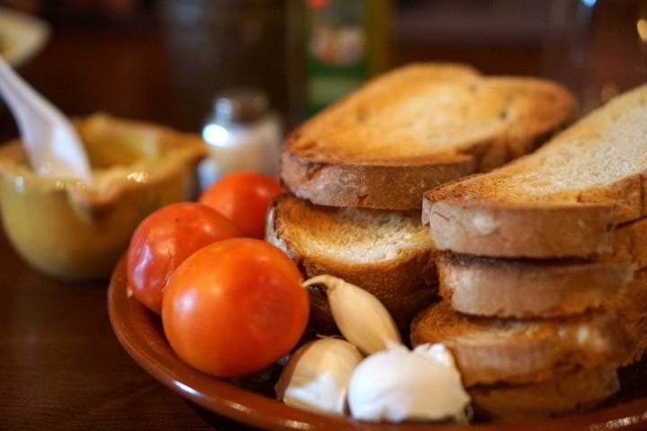 Four photos showing how to make pan con tomate by rubbing garlic and tomato on toast.