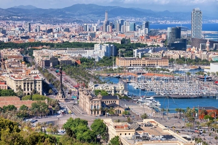 A panoramic view of Barcelona's old buildings, skyscrapers, marina, and mountains in the background.