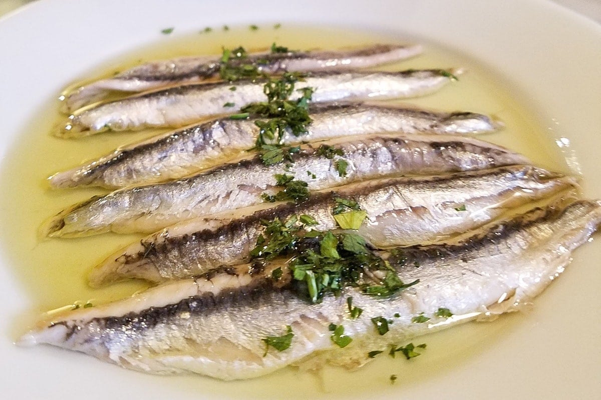 A dish of six anchovies soaked in vinegar and olive oil and topped with fresh herbs.