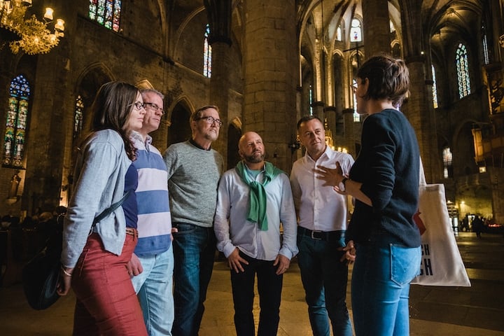 A group of five people listen to a tour guide speaking inside a dimly lit cathedral.