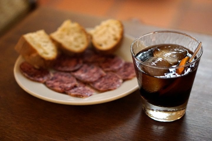 A short glass of vermouth on ice with a plate of cured meat and bread behind it.