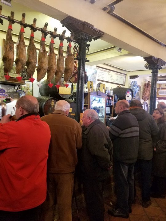 Several older men stand at a bar with legs of cured ham hanging above it