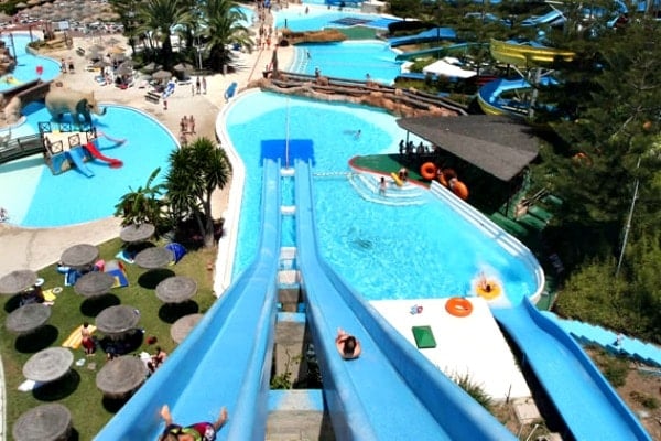 A view from the top of a steep blue waterslide, with several swimming pools below. 