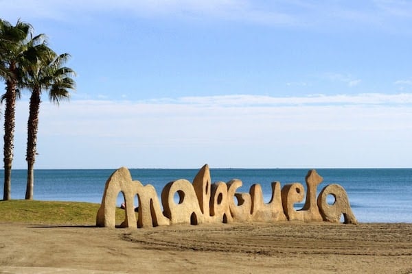 A sand-colored sculpture spelling out "Malagueta" in large letters, with the sea behind it.