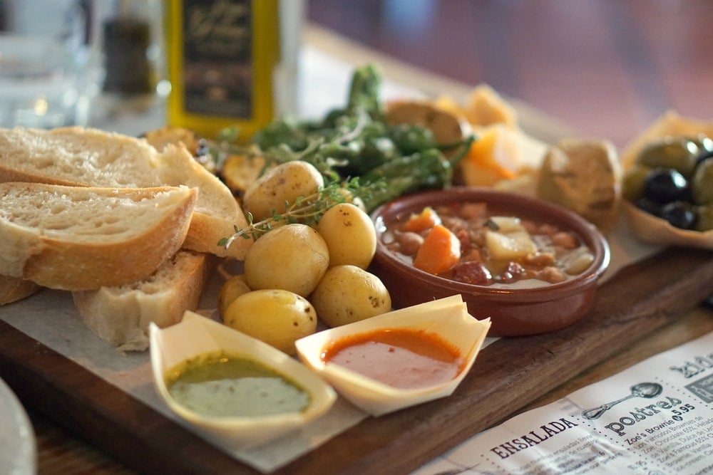 A platter of small potatoes, red and green sauces, bread slices, stew, olives, and other tapas.