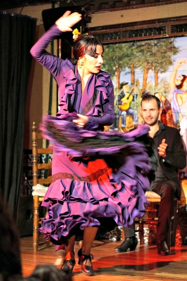 A woman in a purple flamenco dress twirls on stage as another performer claps behind her.