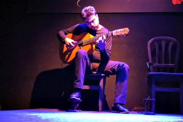 A seated man wearing black plays guitar on a dimly lit stage. 