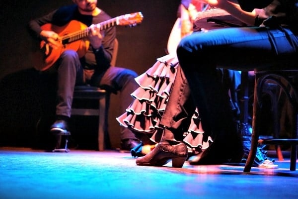 Low angle of three seated flamenco performers on stage, one of whom is playing a guitar.