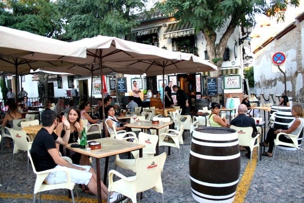 A restaurant terrace with umbrellas, trees, large barrels, and several people sitting at tables with drinks.