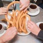 Overhead shot of hands grabbing churros from a plate with three mugs of chocolate