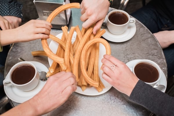 Overhead shot of hands grabbing churros from a plate with three mugs of chocolate