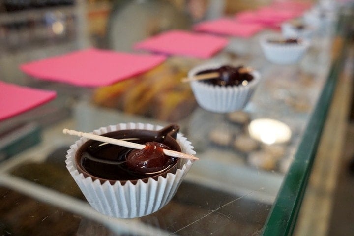 Small paper liners containing chocolate cups of dark liquid with skewered cherries on a glass countertop.