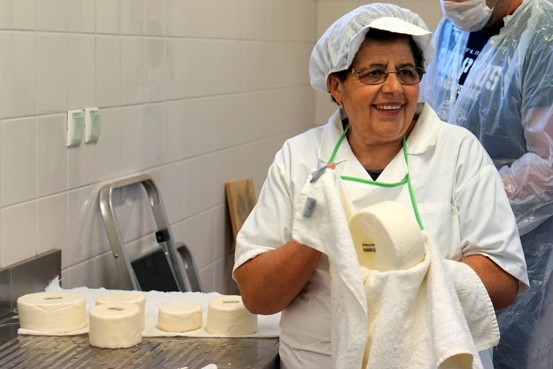 A woman wearing a hairnet holds a wheel of white cheese in a towel.