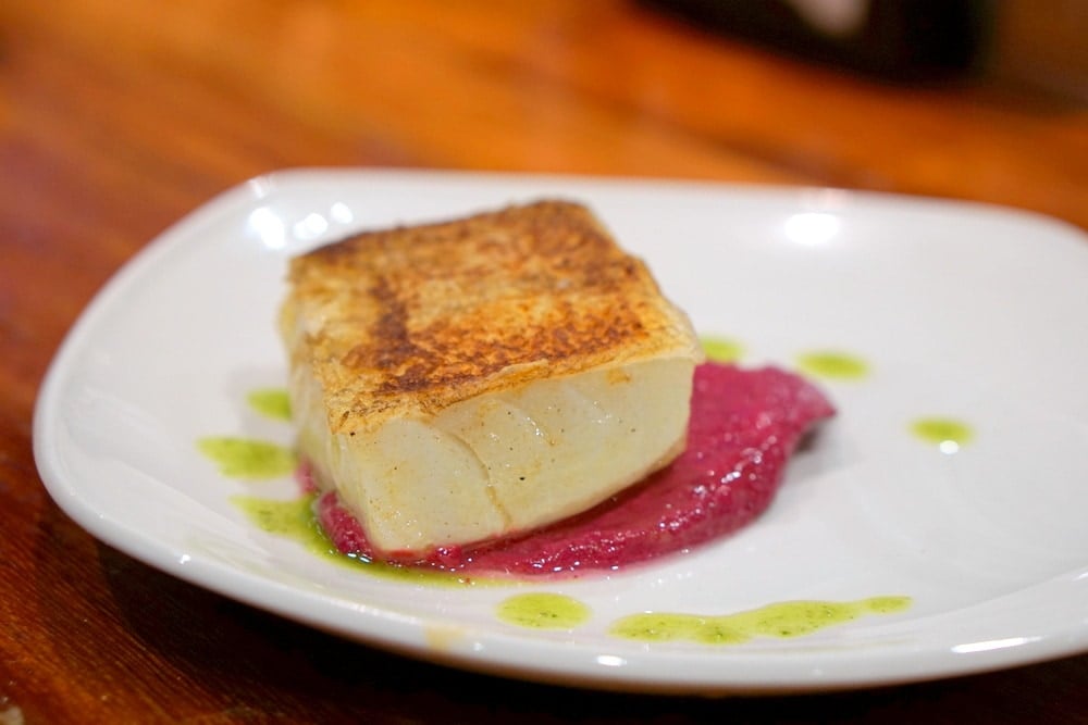 A piece of golden-brown salt cod with pink sauce and green oil on a white plate