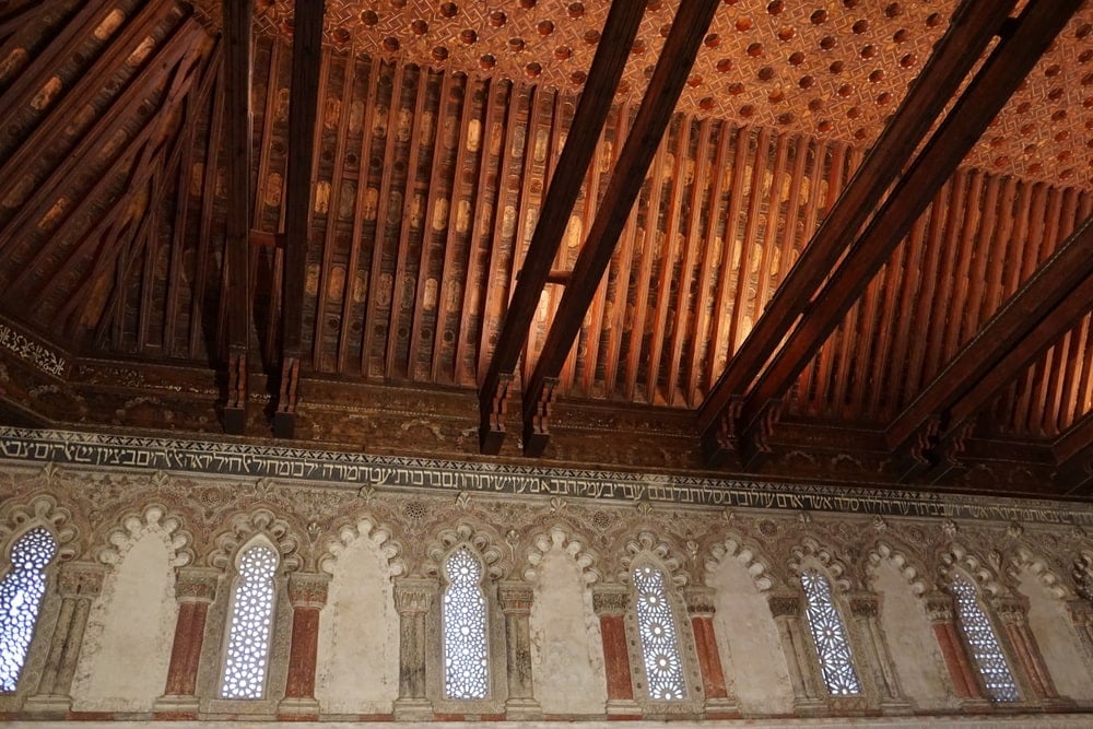 An intricate wooden ceiling and walls with ornate arched windows and Arabic writing above them.