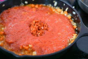 Tomato puree and chopped chorizo on top of fried onions and carrots in a skillet