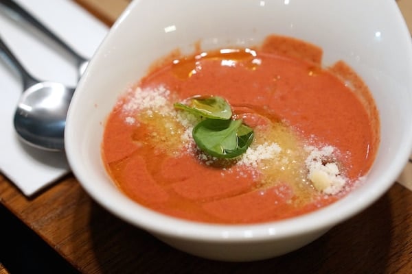 A bowl of bright orange soup topped with basil leaves, parmesan cheese, and olive oil.