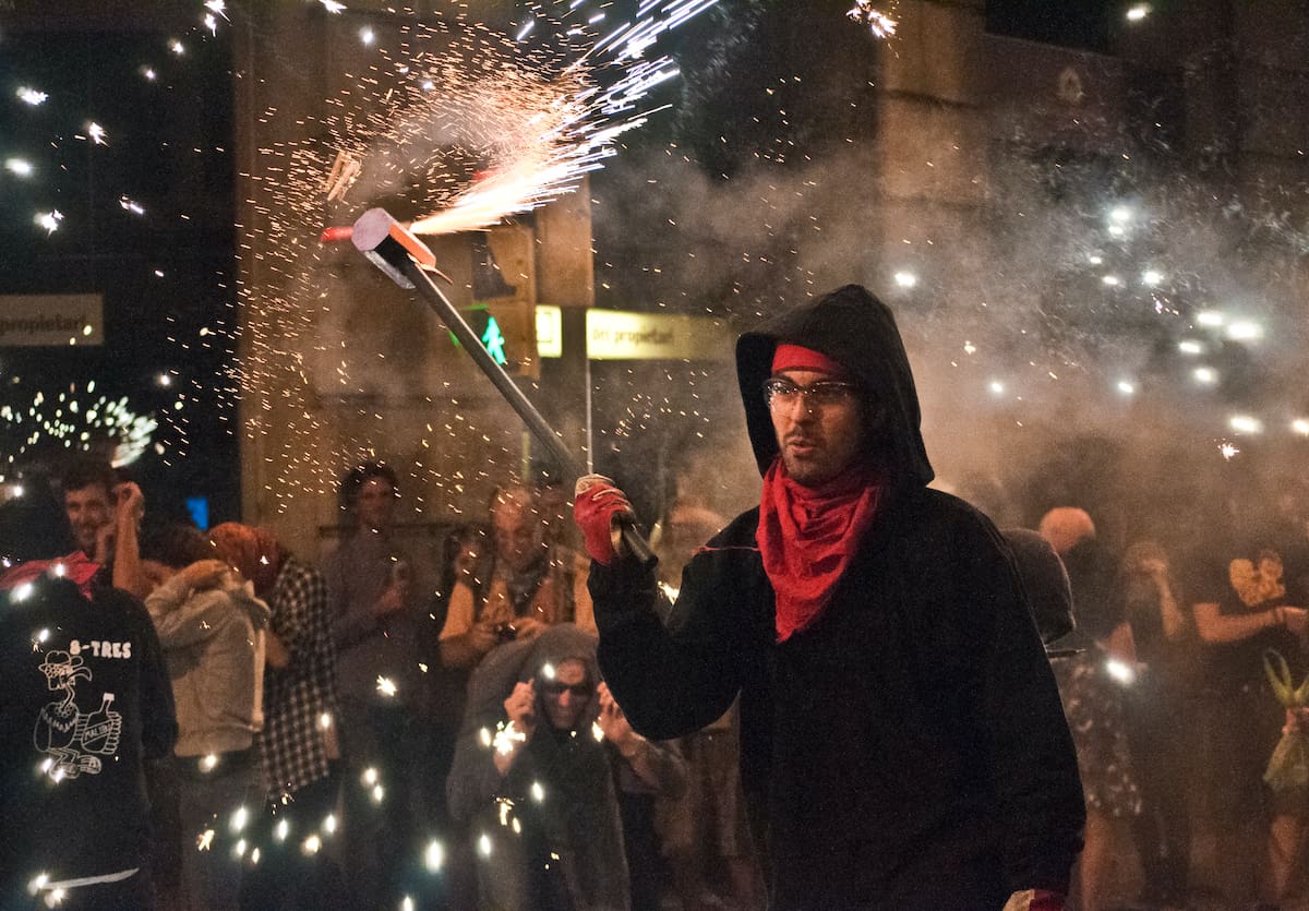 A person in a hoodie on a crowded street in Barcelona holding a festive flame-throwing device. 