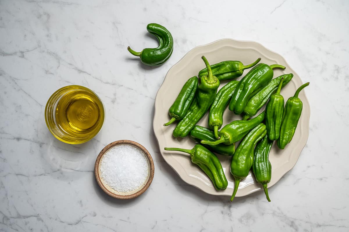 Ingredients for making padron peppers on a white marble counter. Cup of olive oil, sea salt and green padron peppers.