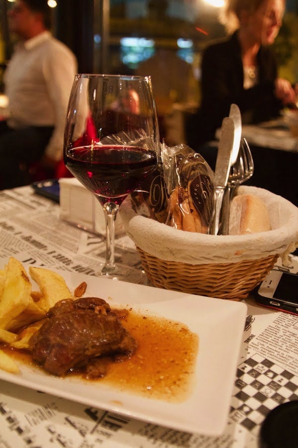 Table with a bread basket, glass of red wine, and plate of stewed meat and potatoes.