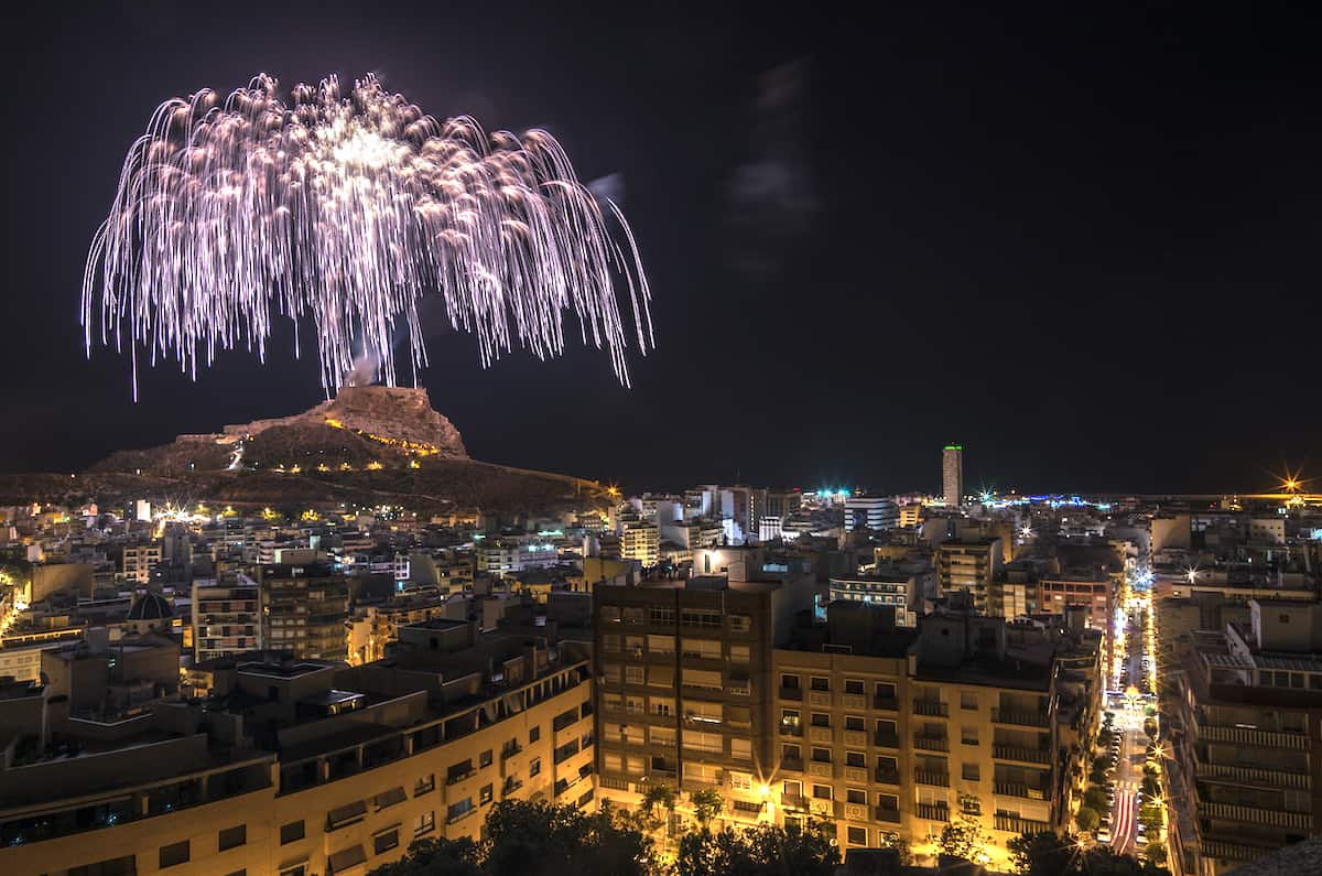 A wide view of Alicante at night with silver fireworks lighting up the sky.