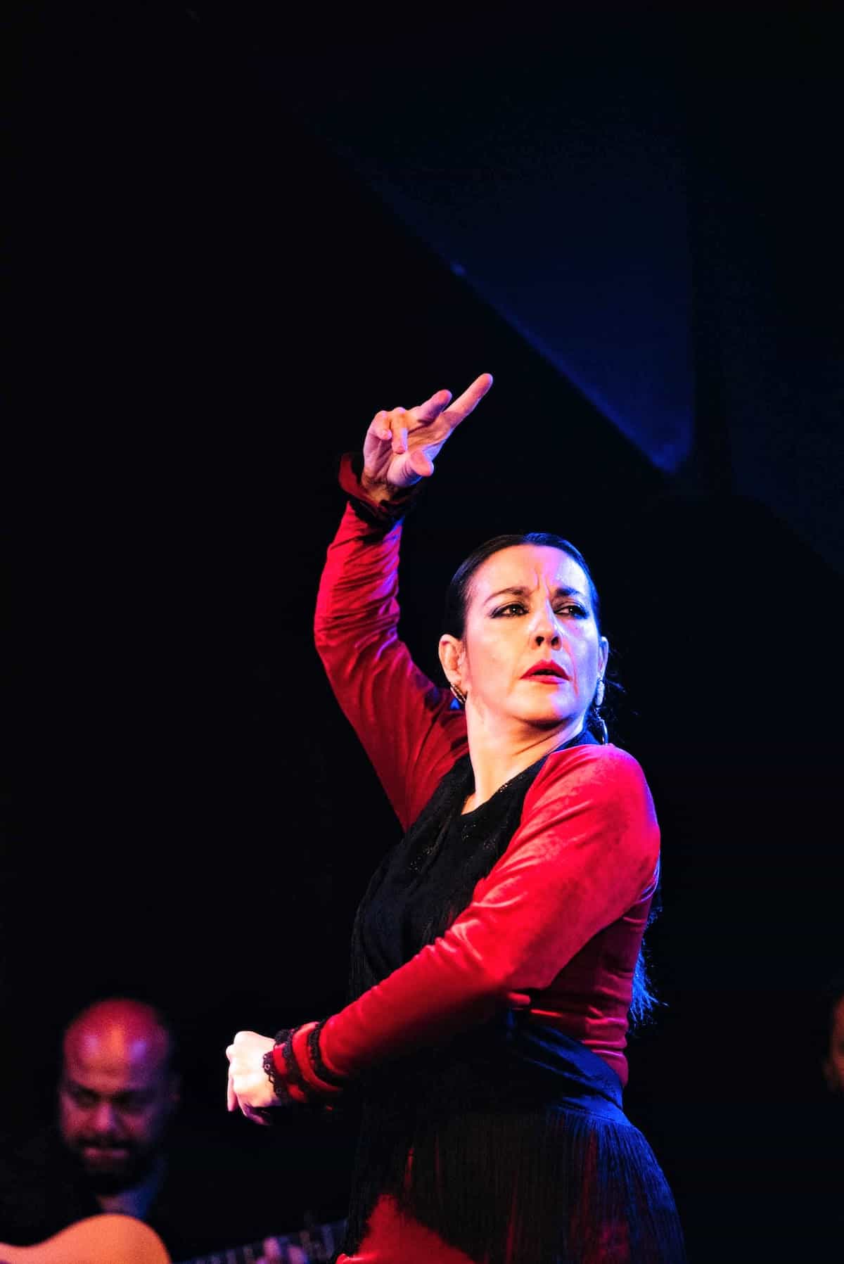 A flamenco dancer dressed in red and black performing with a serious look on her face.