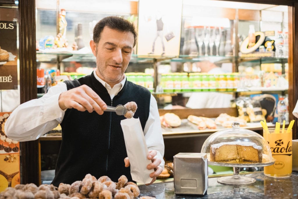 Man using tongs to put a sugary doughnut into a paper bag at a pastry shop.