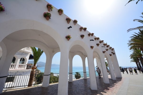 A white pergola with arches and potted flowers on the sides, with the sea behind it.