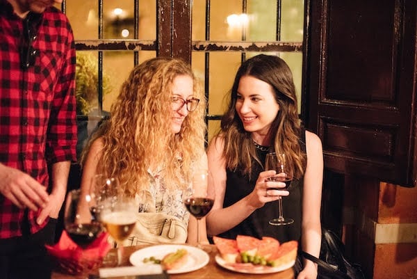 Two smiling women sit at a small restaurant table with glasses of wine and tapas. 