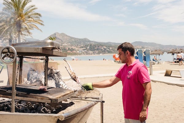 A man places a skewered sardine over a smoking grill on a sandy beach.
