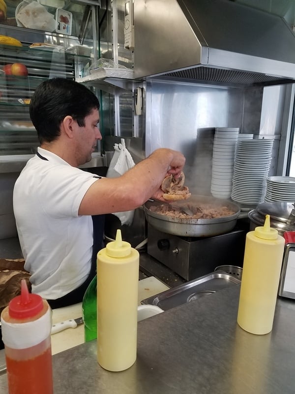 A cook assembles a bifana, placing stewed pork from a large pot onto a bread roll.