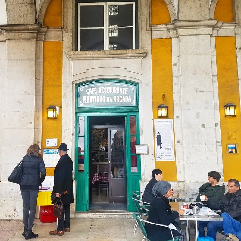 An old building with yellow walls and a green doorframe, with people sitting at tables outside.